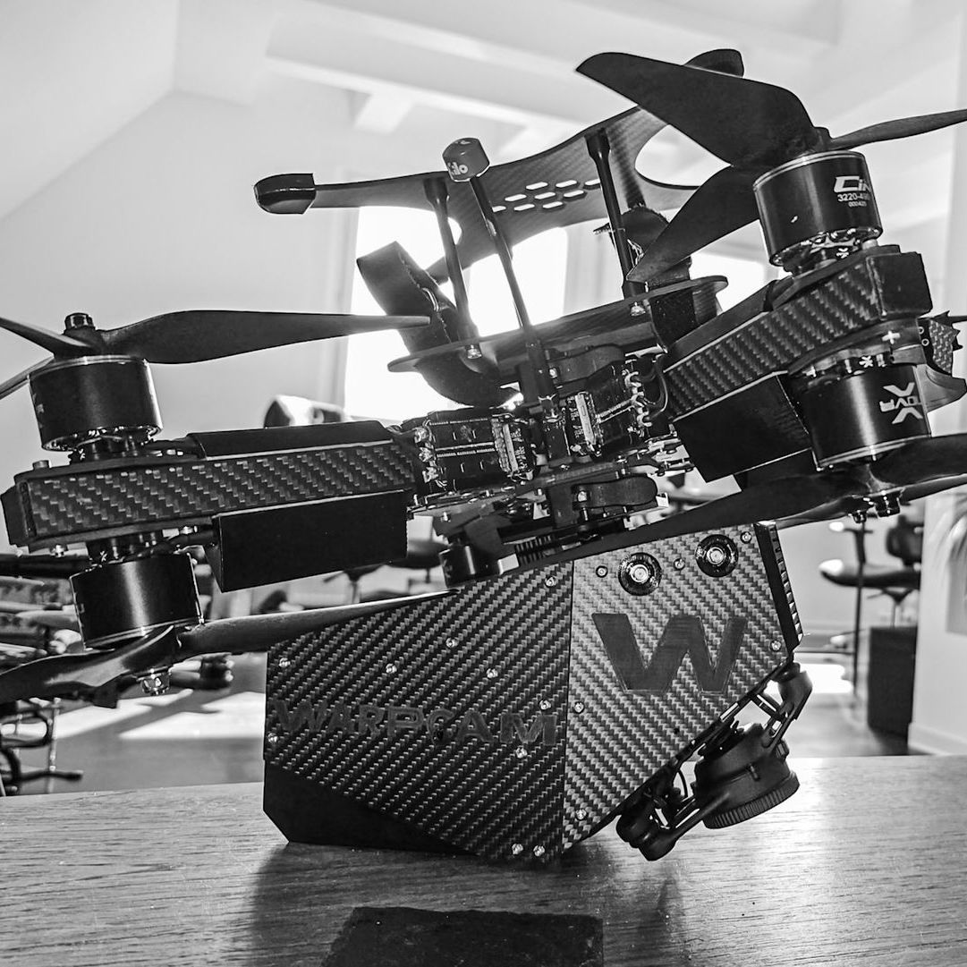 WarpCam unit without gimbal attached to a carbon fiber FPV racing drone, lying on a table between shots, showing the compact lightweight rig used for high speed low angle cinematic footage.
