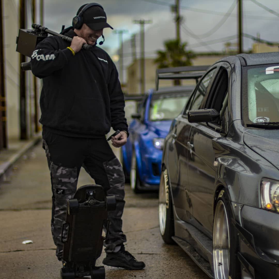 Director Ferdi Fischer on an Amazon Prime car show set, standing with an early WarpCam prototype slung over his shoulder while briefing the drivers of two heavily modified, Fast and Furious style cars, his electric skateboard at his feet on the tarmac as they prepare for the next dynamic driving shot.