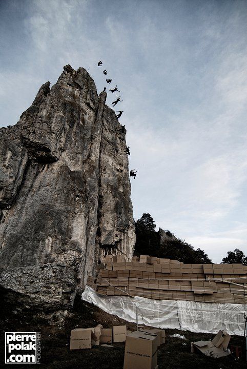Side view composite photo showing multiple positions of Ferdi Fischer performing a backward gainer from the 45 meter Burgsteinfelsen during his world record high fall into stacked cardboard boxes, the entire arc of his descent captured step by step in one image with the landing boxes and spare boxes visible on the ground.