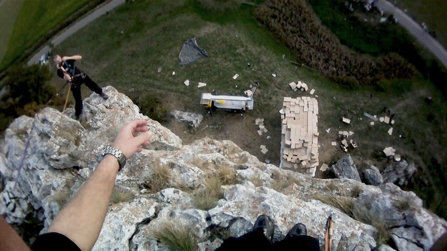 First person GoPro view from Ferdi Fischer standing on top of the 45 meter Burgsteinfelsen in Bavaria just before his world record high fall for the TV show Galileo, with his arms and feet visible at the edge of the rock and dozens of cardboard boxes far below appearing tiny in the distance.