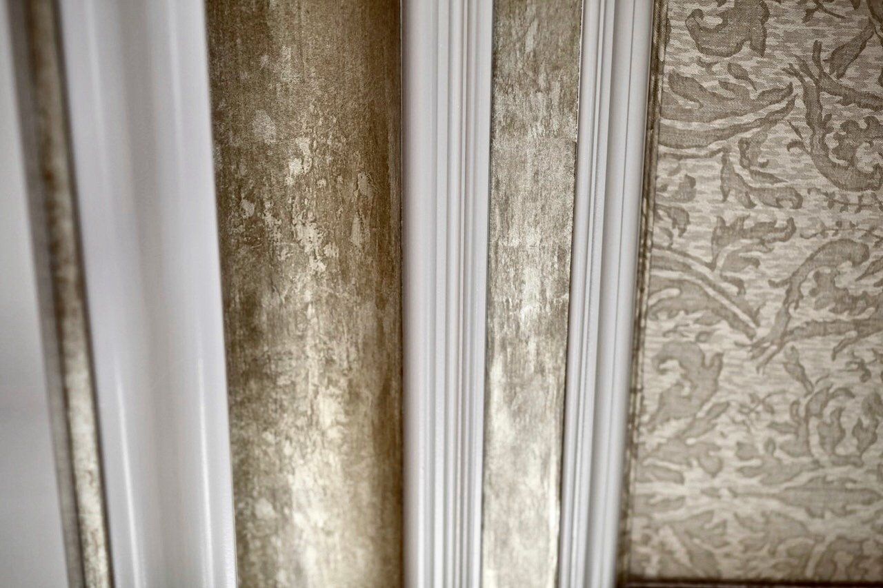 A close up of a wall with a white trim and gold wallpaper.