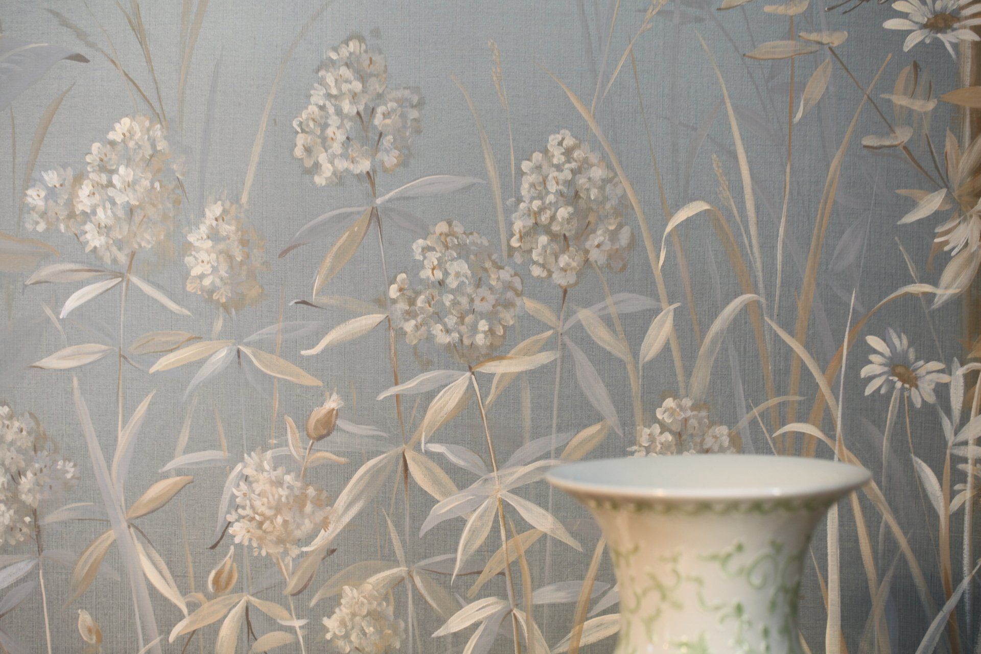 A vase is sitting in front of a painting of flowers on a wall.