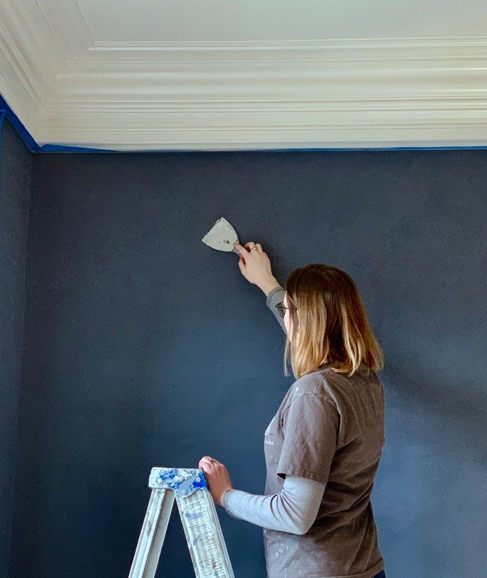 A woman is standing on a ladder painting a wall with a spatula.