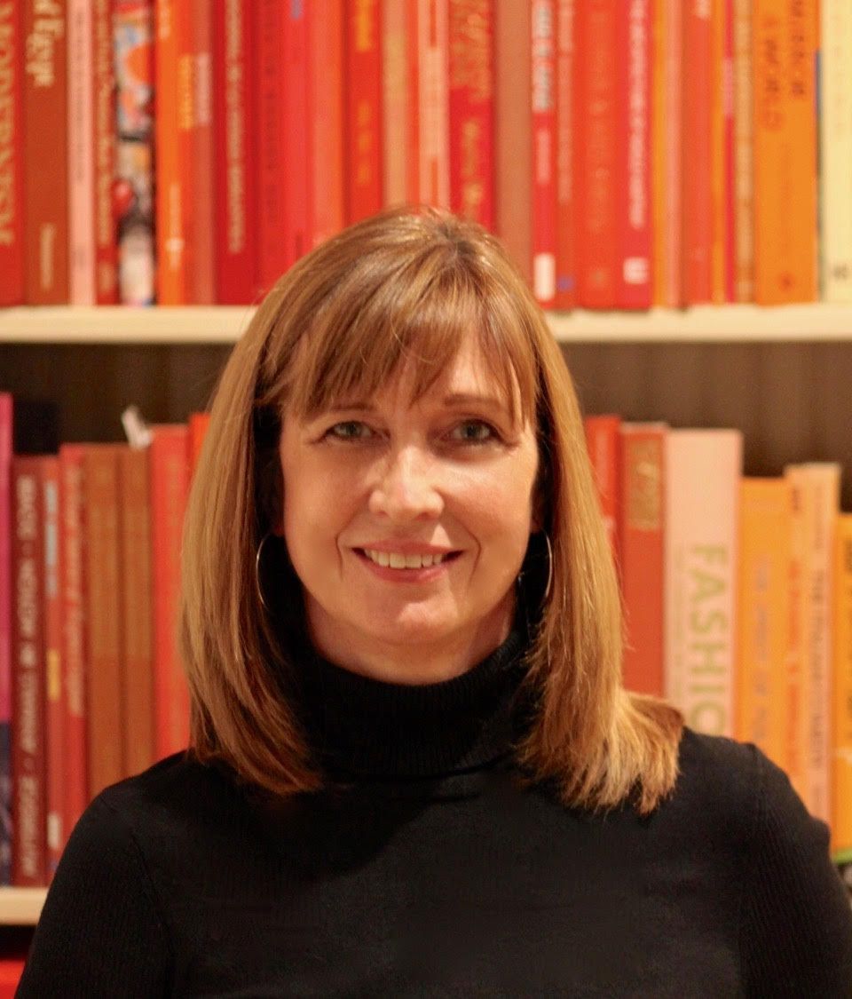 A woman in a black turtleneck is smiling in front of a bookshelf full of books