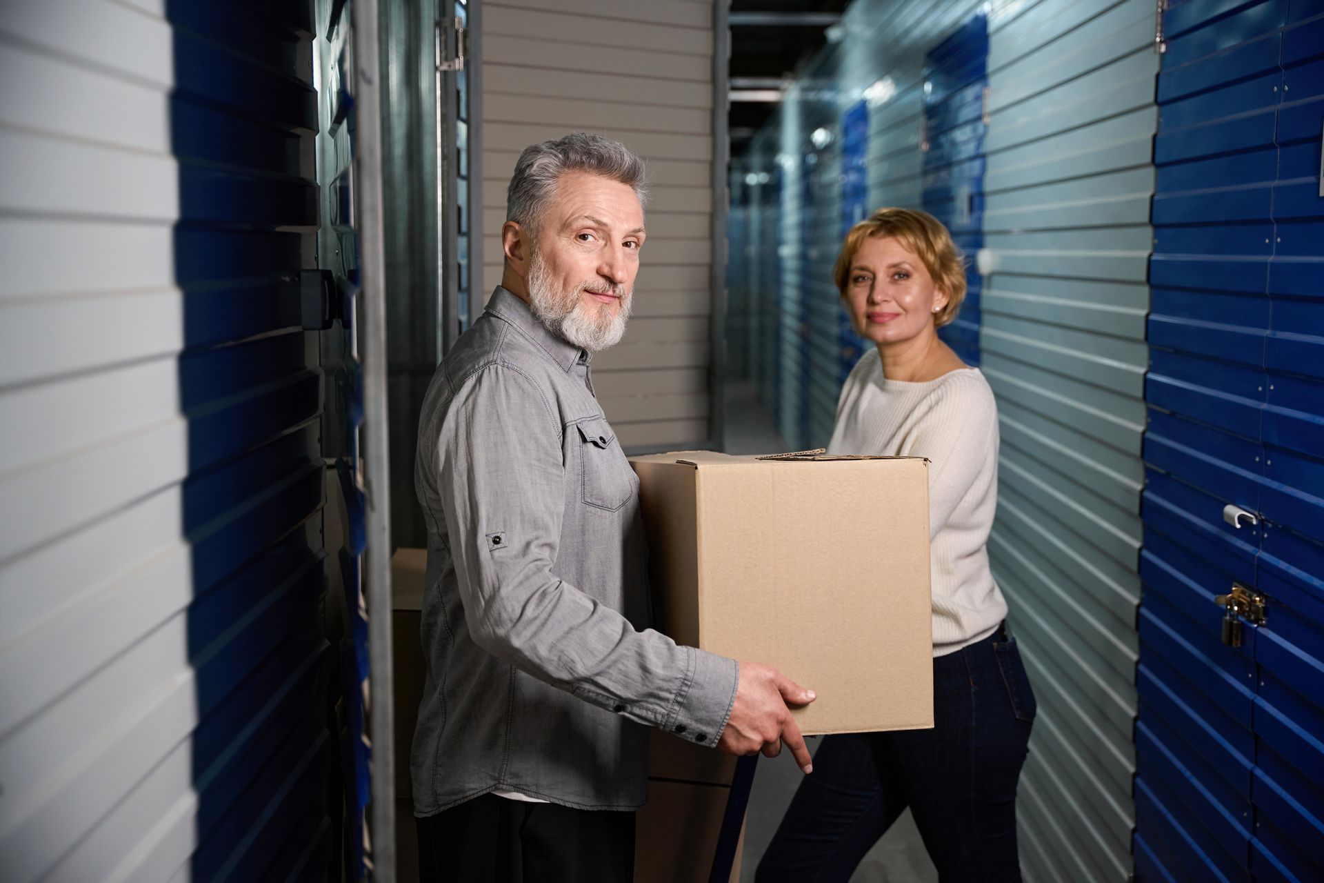 Smiling couple carrying a box inside a storage facility hallway with blue doors.