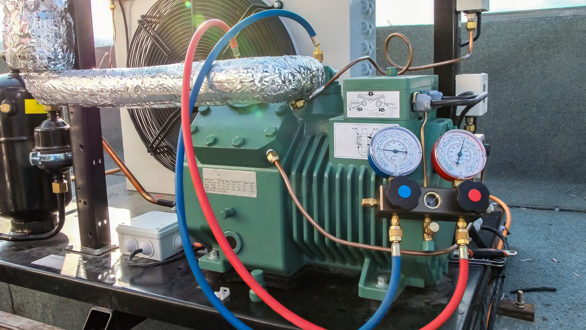 Green refrigeration unit with blue and red hoses attached, gauges, and insulated pipes, outdoors.