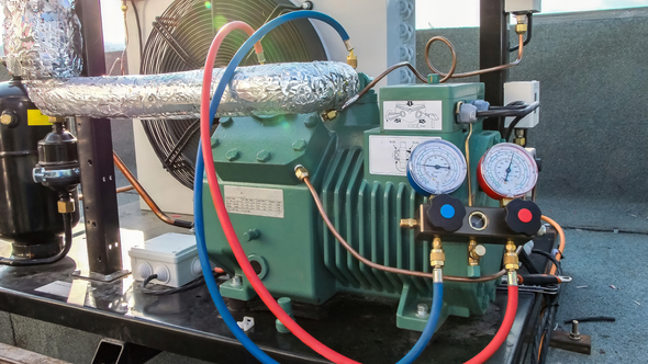 Green refrigeration unit with blue and red hoses attached, gauges, and insulated pipes, outdoors.