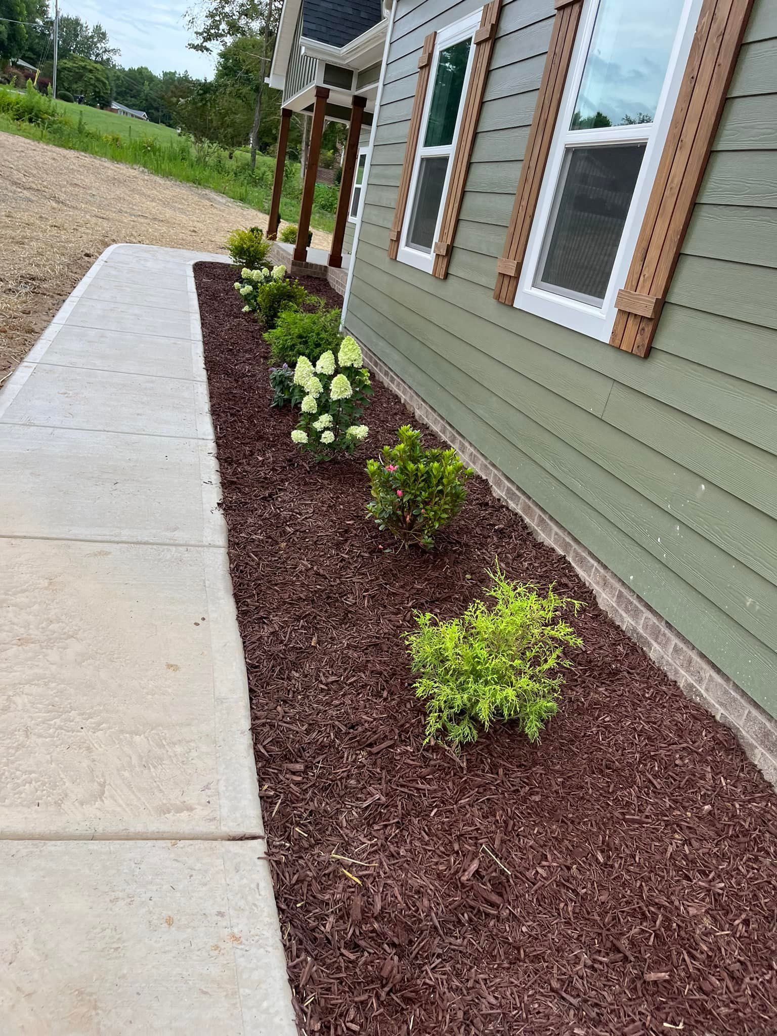 A narrow flowerbed with plants, along a sidewalk and the side of a green house.