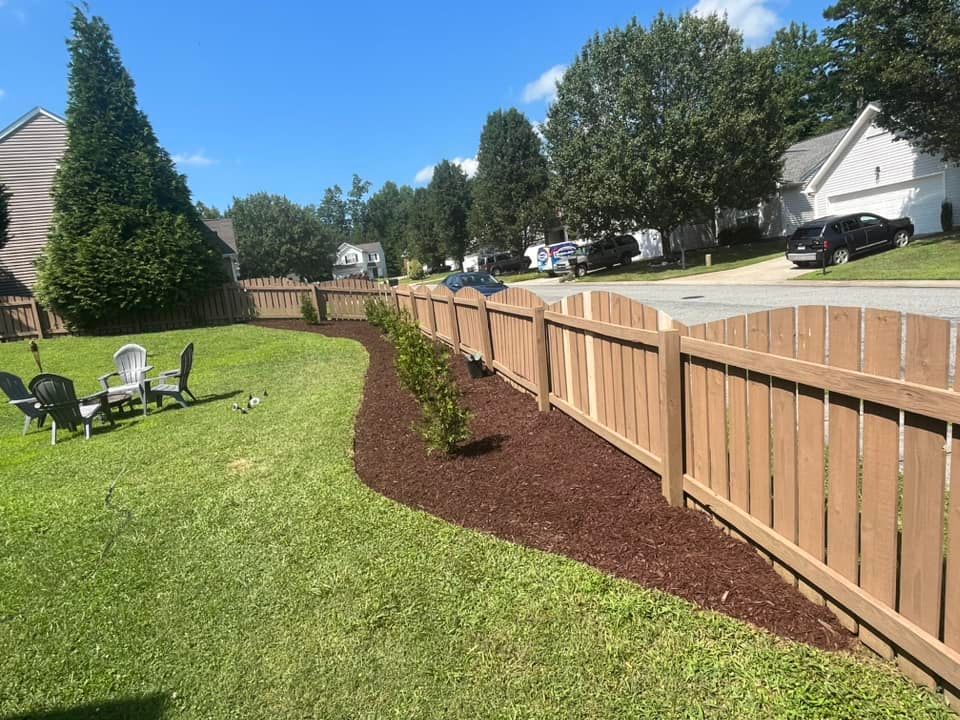 Brown wooden fence borders a grassy yard with fresh mulch and small shrubs. Houses line the sunny street.