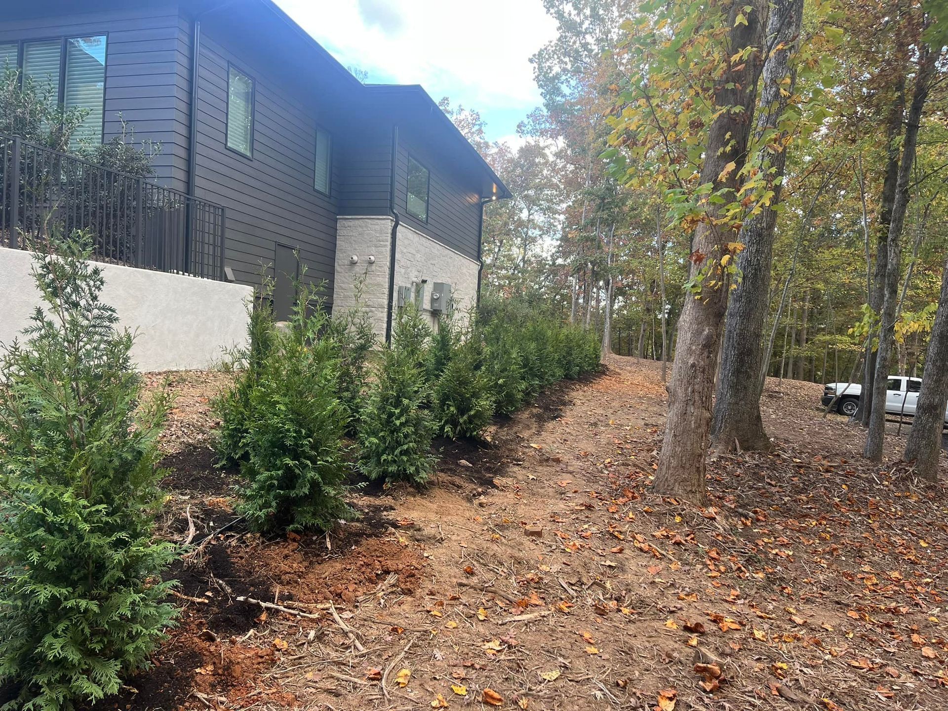 A house with dark siding and a stone base next to a dirt path with bushes and trees.