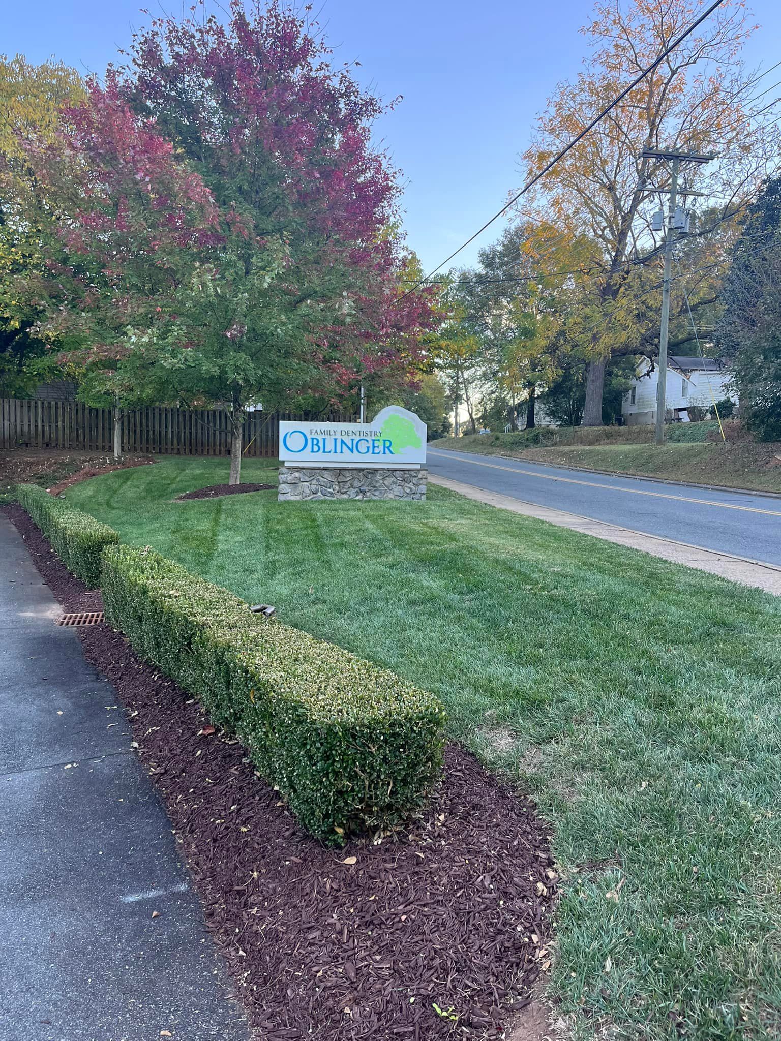 A park sign on a grassy lawn with trimmed hedges in front; trees with colorful autumn leaves are behind it.