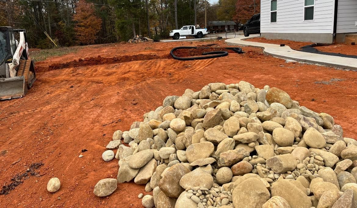 Pile of rocks in front of red dirt, a construction site, with a trench and house in the background.