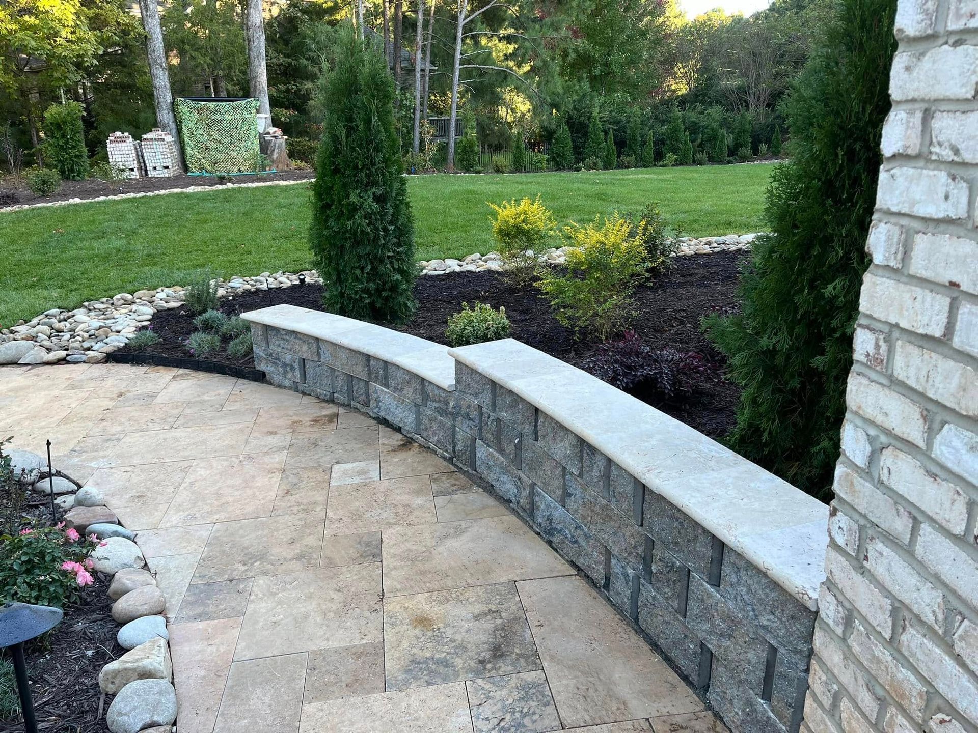 Stone walkway curving toward a raised garden bed with a stone wall and evergreen trees.