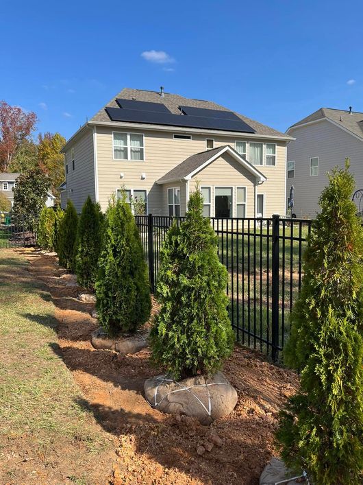 Backyard with a two-story house, solar panels, green shrubs, black fence, and a blue sky.