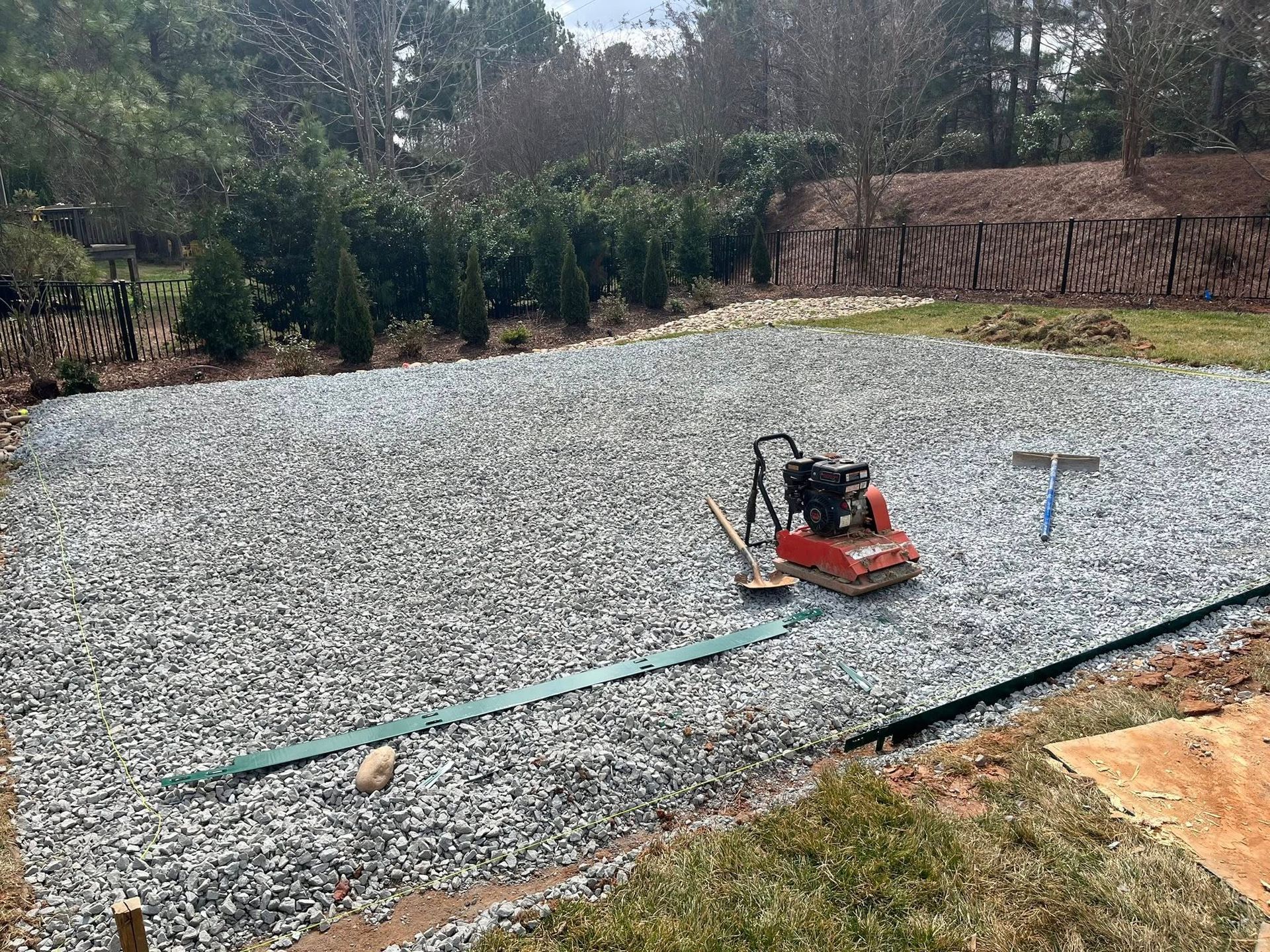 Gravel-covered yard, prepared for a structure, with a compactor and rake; surrounded by greenery.
