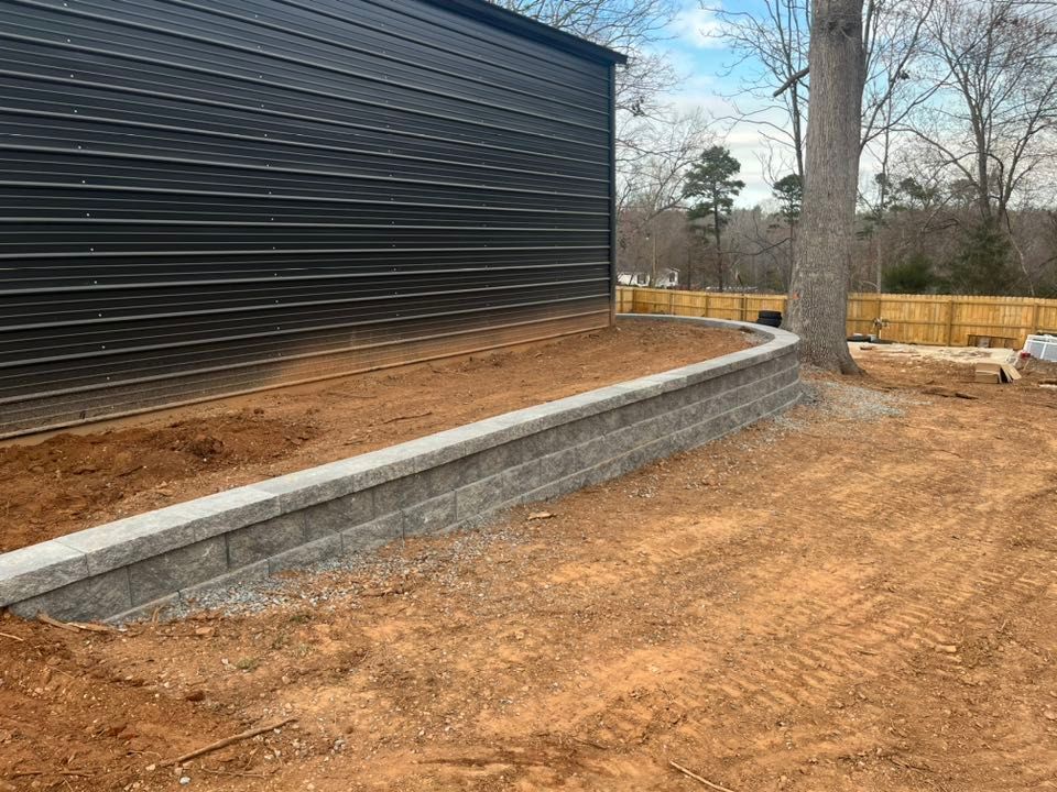 Retaining wall with gray blocks next to a black building and a tree, in a yard with dirt.