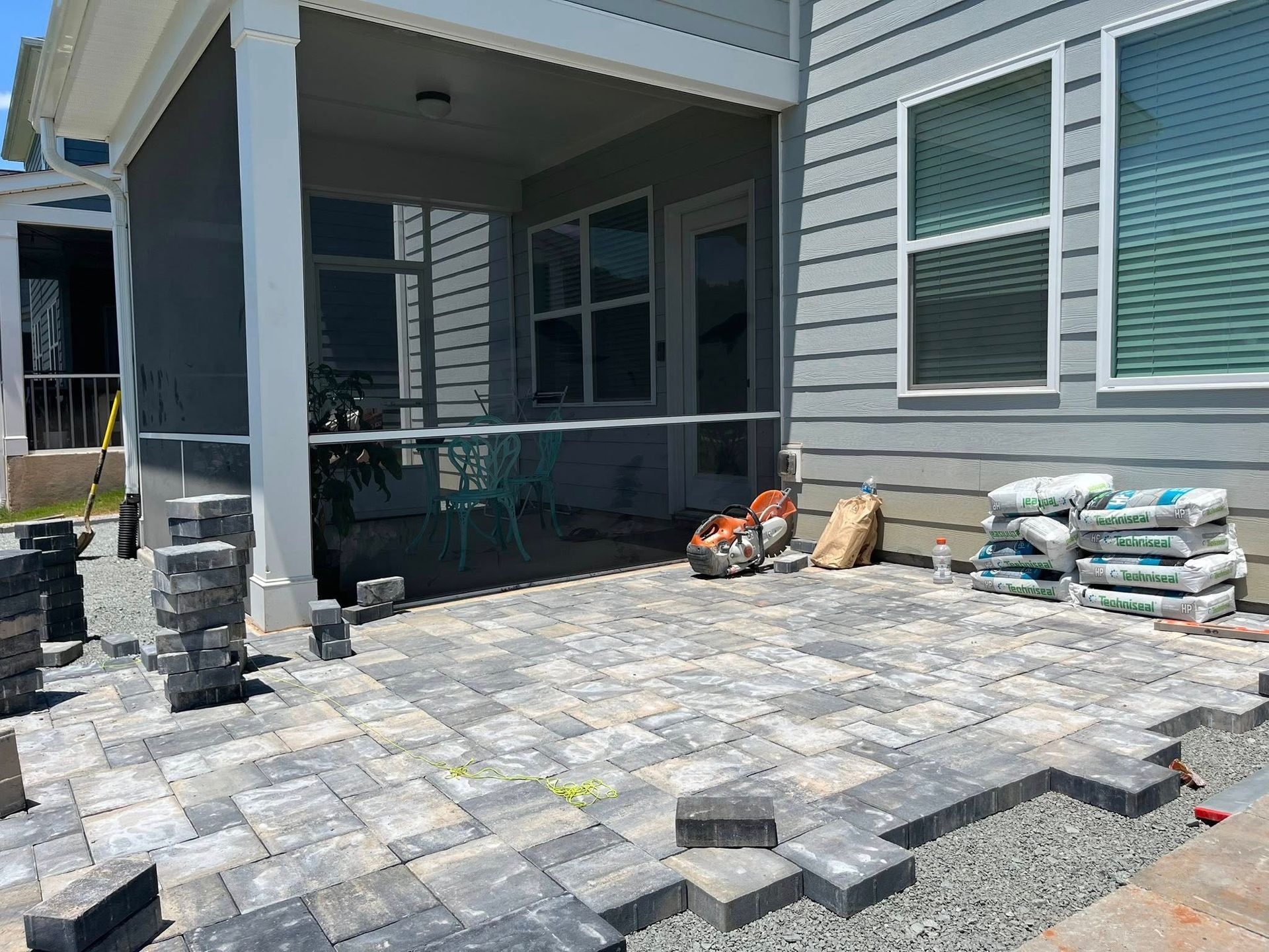 Patio being constructed with gray and tan pavers, near a light gray house. Bags of materials are present.