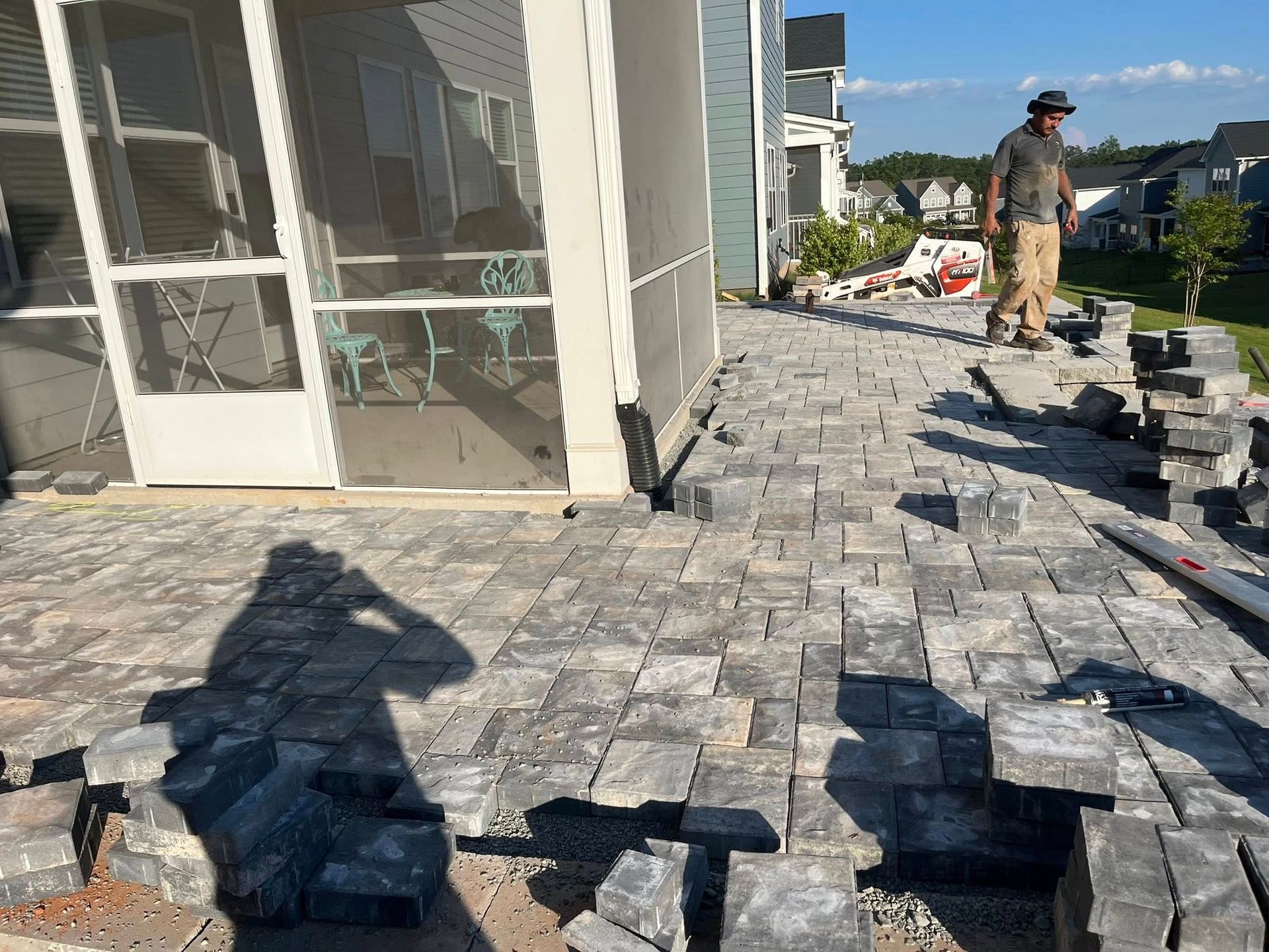 A man lays grey pavers on a patio next to a screened porch in a suburban setting.