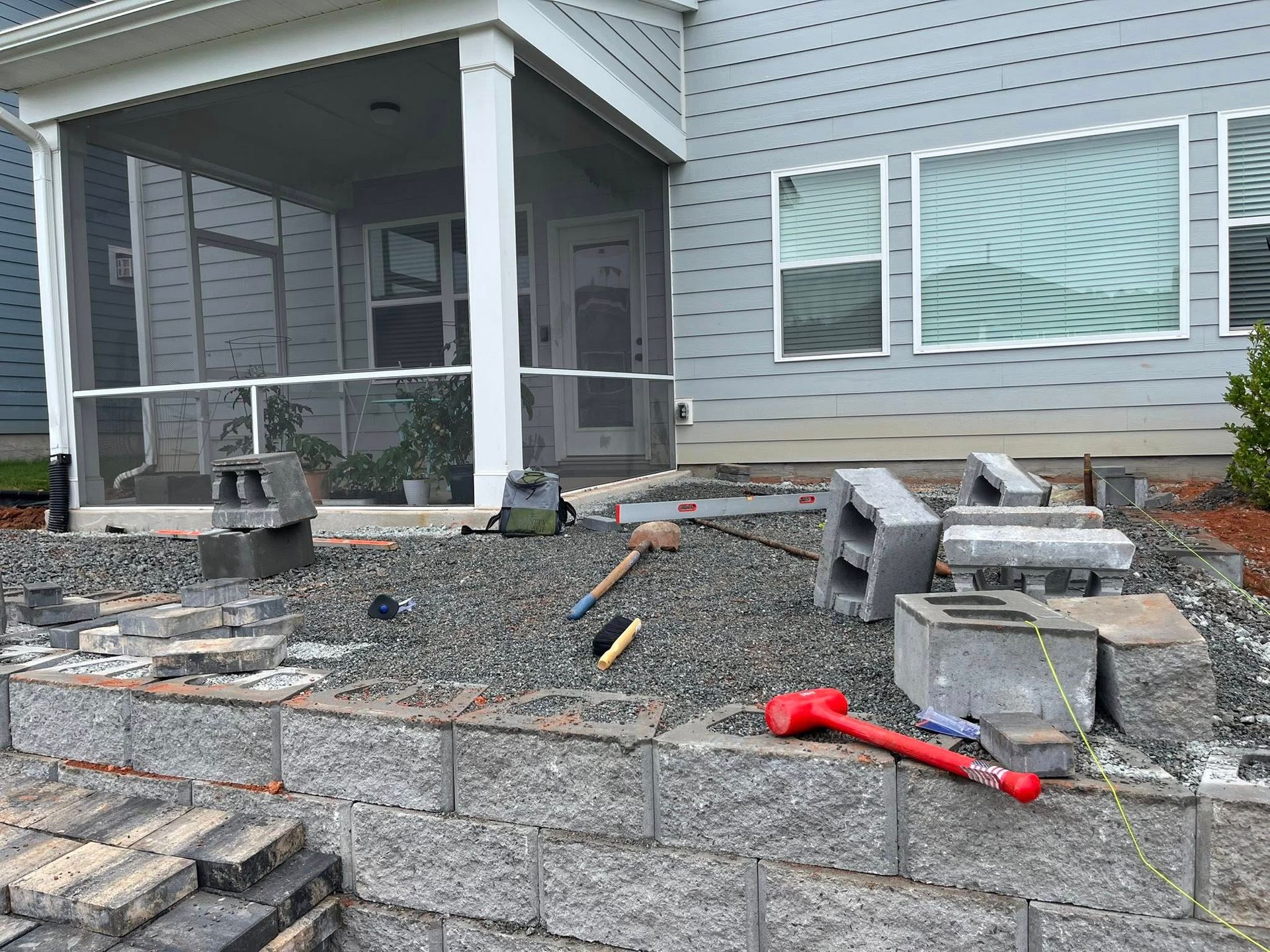 Construction site with retaining wall, gravel, and cinder blocks near a house with a screened porch.