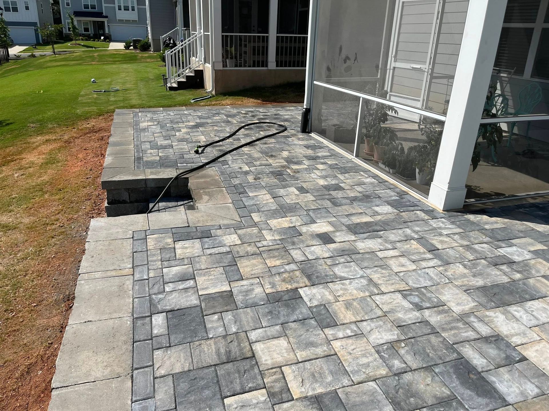 Patio with patterned gray pavers next to a screened porch, with a hose across the surface.