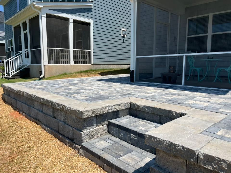 Stone patio with steps and retaining wall, adjacent to a house with screened porch.