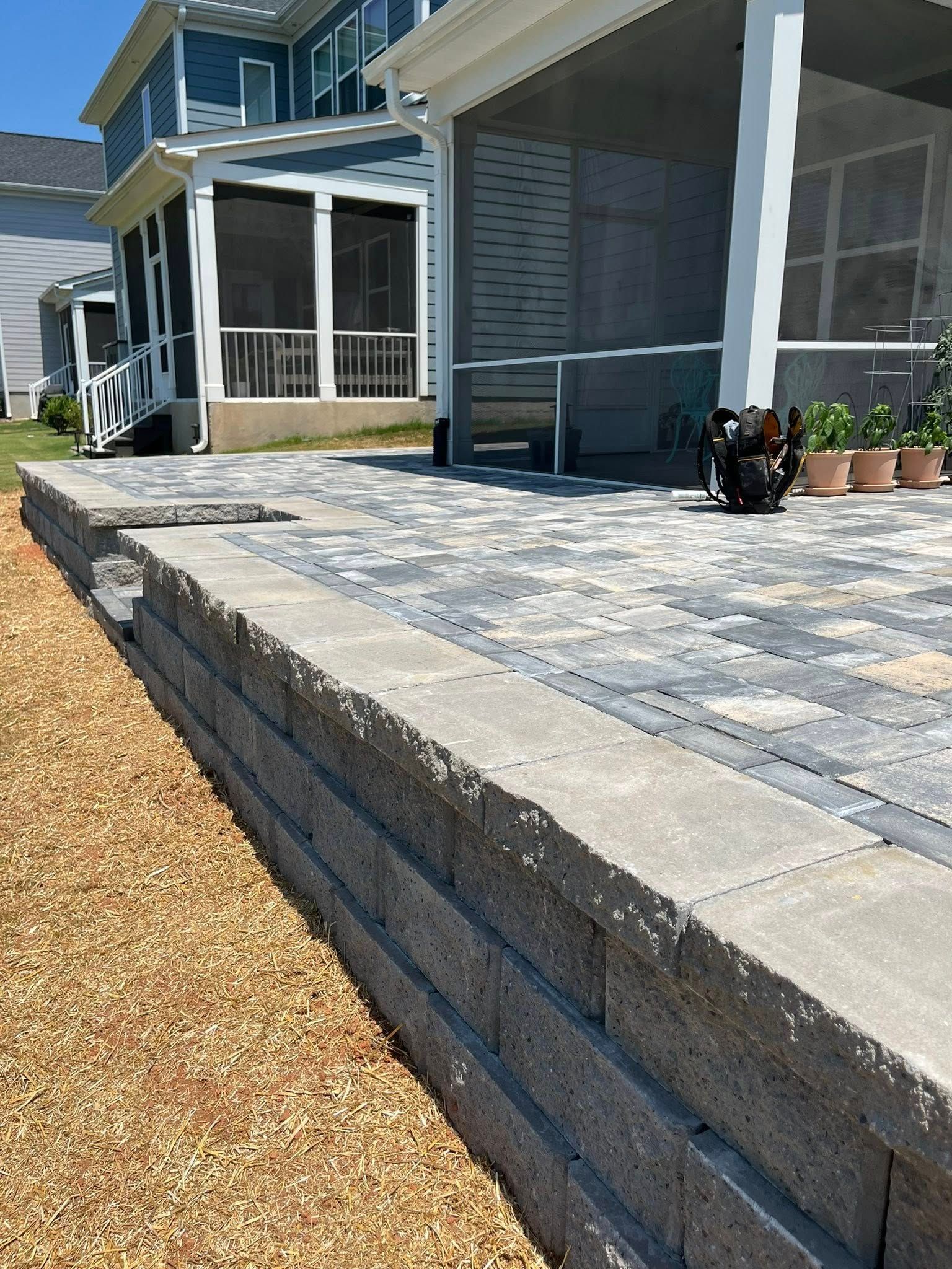 Stone retaining wall bordering a patio with patterned pavers, next to a house with screened porch.