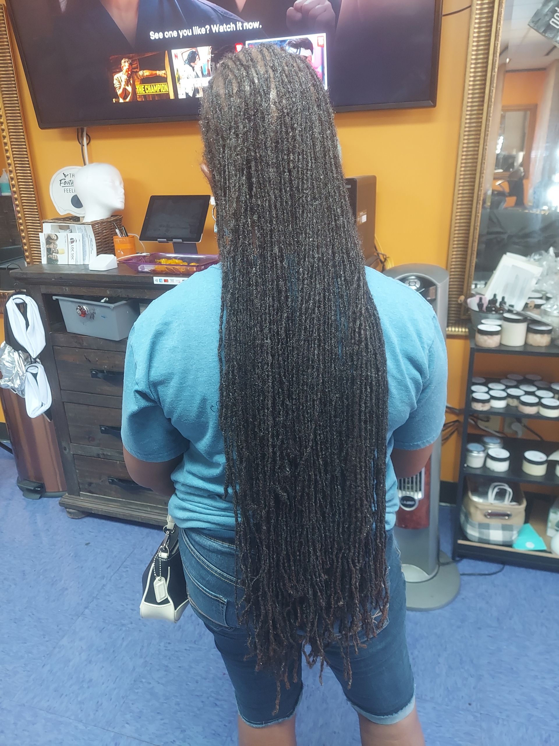 Person with long, black braids stands in a salon. They wear a blue shirt and denim shorts.