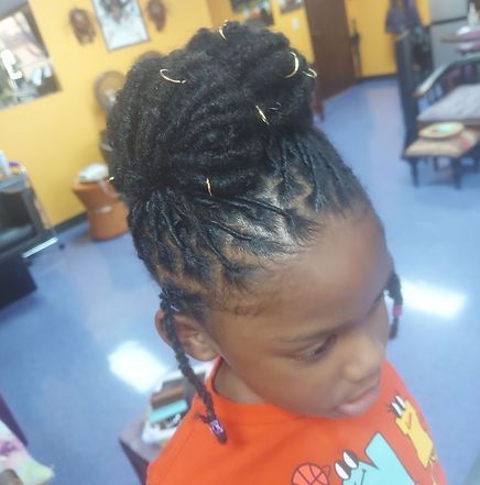 Young Black child with locs styled in a high bun, wearing an orange shirt, indoors.