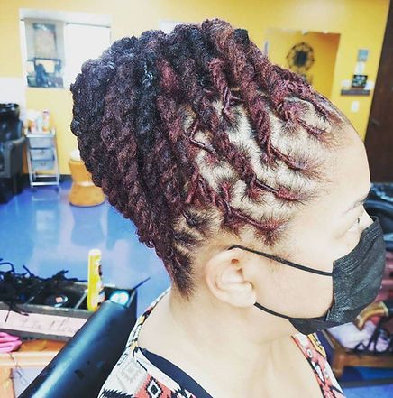 Woman with burgundy locs styled up, wearing a black mask, in a salon with blue floor.