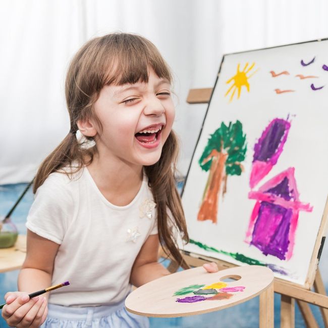 A little girl is laughing while painting on an easel