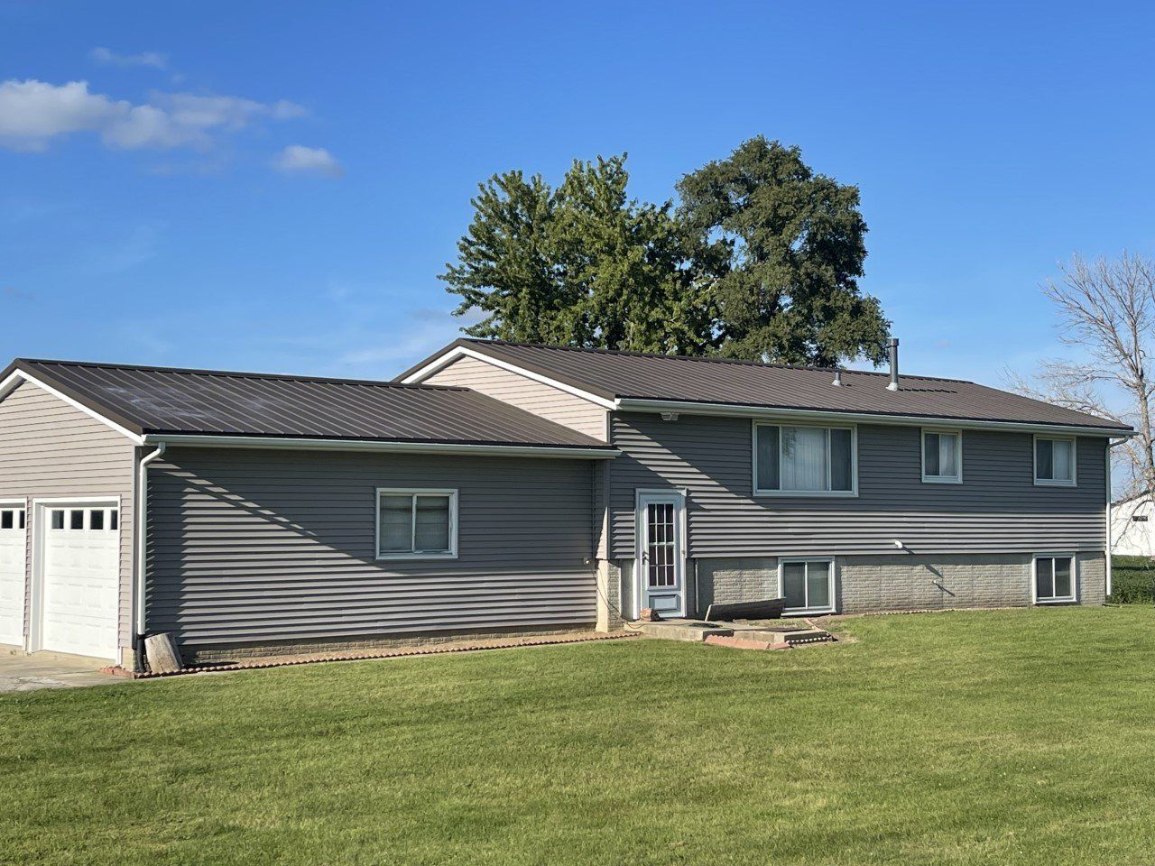 Gray-sided house with a garage, gray roof, and green lawn under a blue sky with trees.
