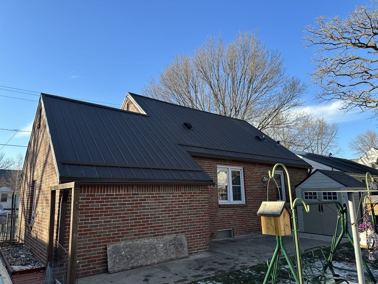 Brick house with dark metal roof on a sunny day, backyard with birdhouse, swing set, and trees.