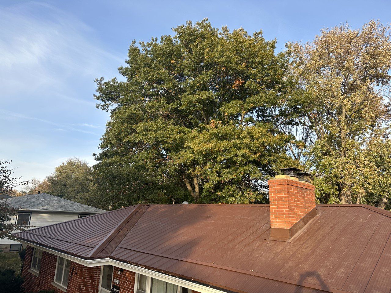 Red brick house with brown roof, chimney, and large leafy trees against a blue sky.