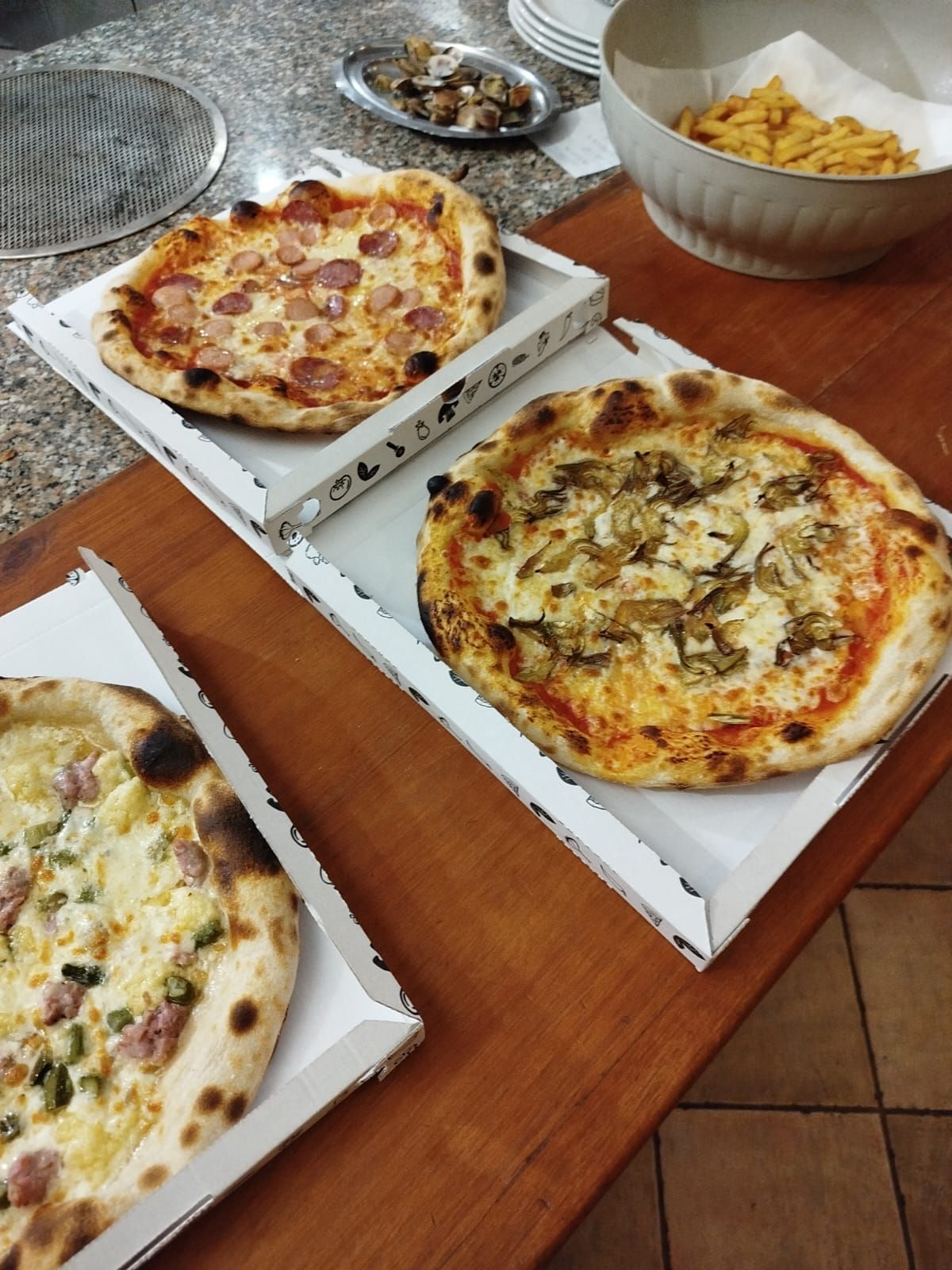 Diverse pizze in scatole aperte su un tavolo di legno, con una ciotola di patatine fritte sullo sfondo.