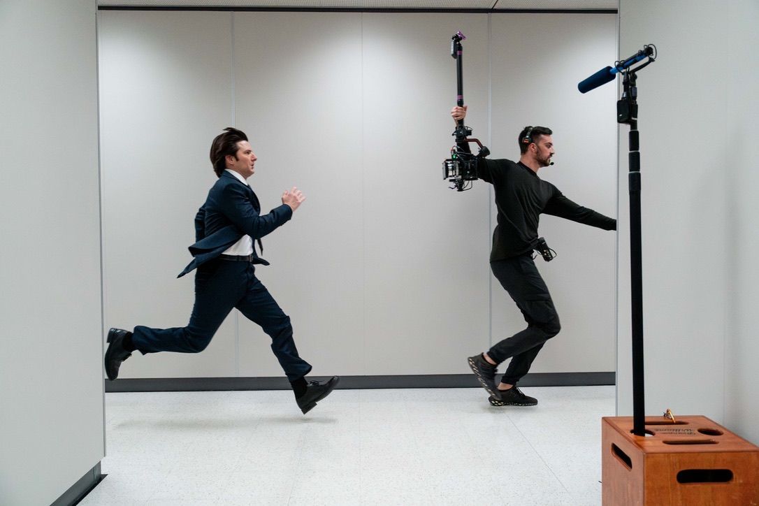 Two men are running in a room and one is holding a camera.