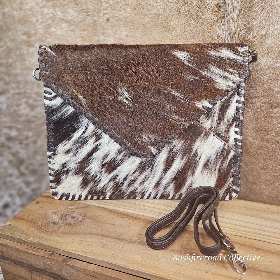 Brown And White Cowhide Clutch With A Brown Leather Strap, On A Wooden Surface — Bushfire Road Collective in Mareeba ,QLD