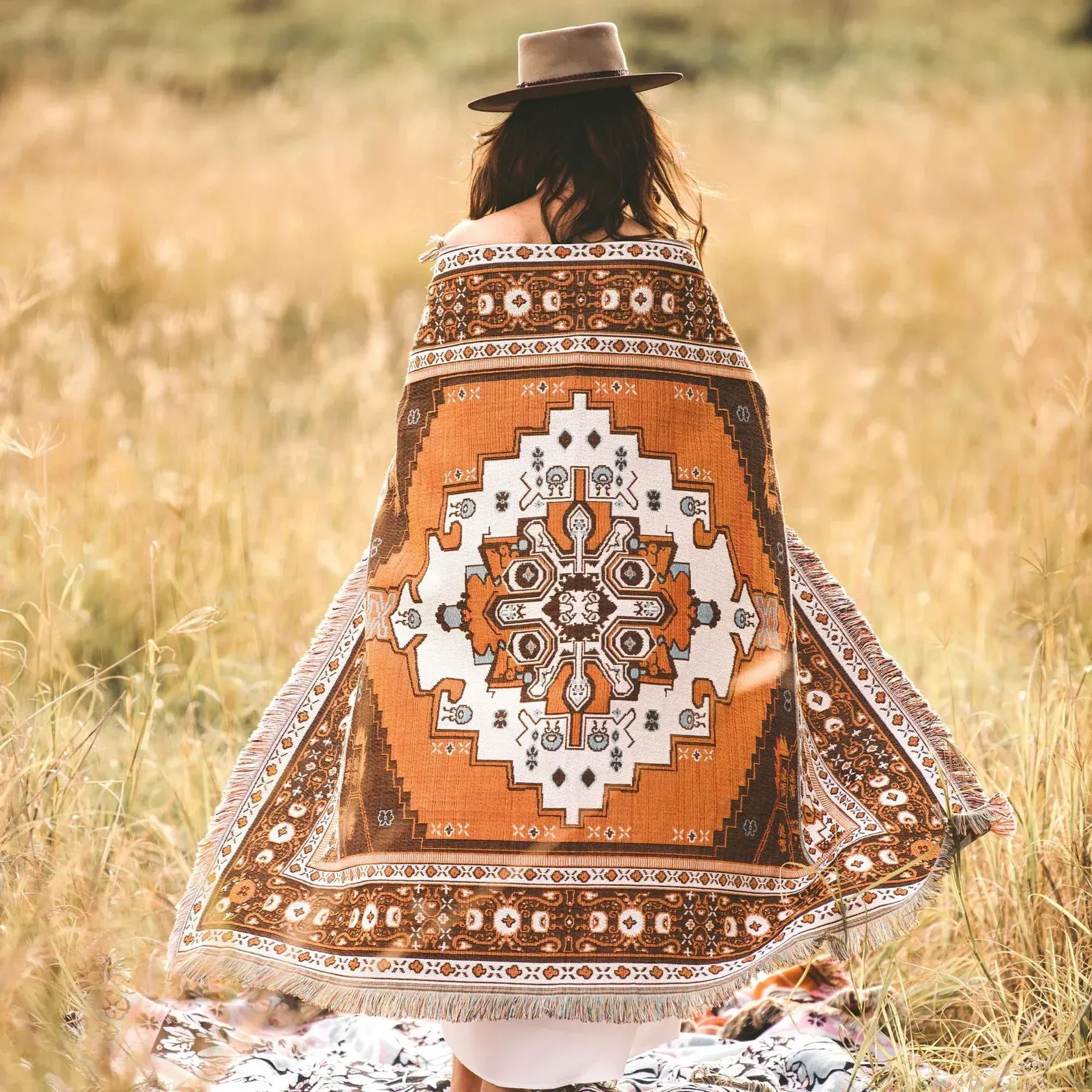 Woman In A Hat, Back To Viewer, Wrapped In A Patterned Orange And Brown Blanket In A Field — Bushfire Road Collective in Mareeba ,QLD