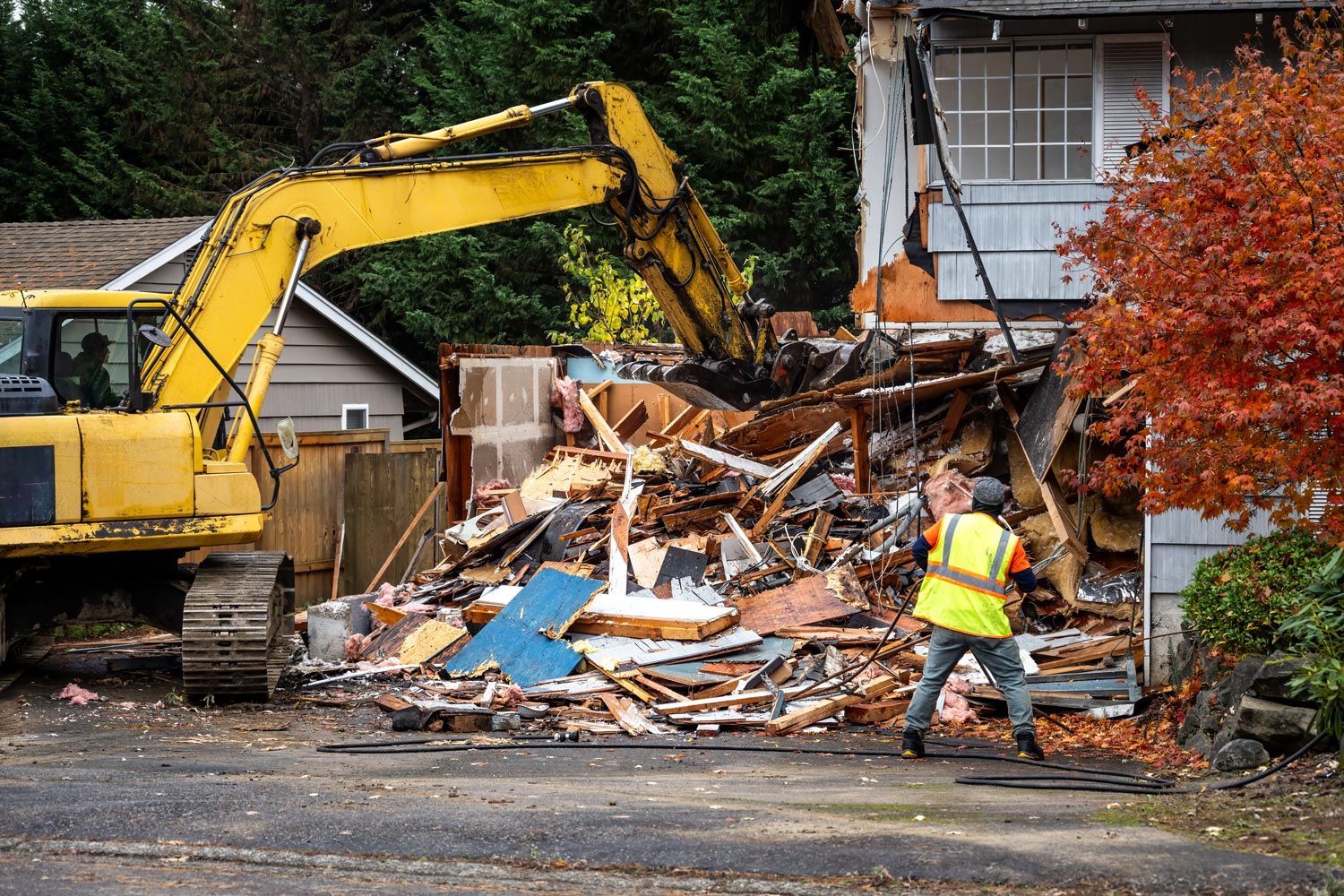 Yellow excavator demolishing a house as a worker in a reflective vest stands beside rubble