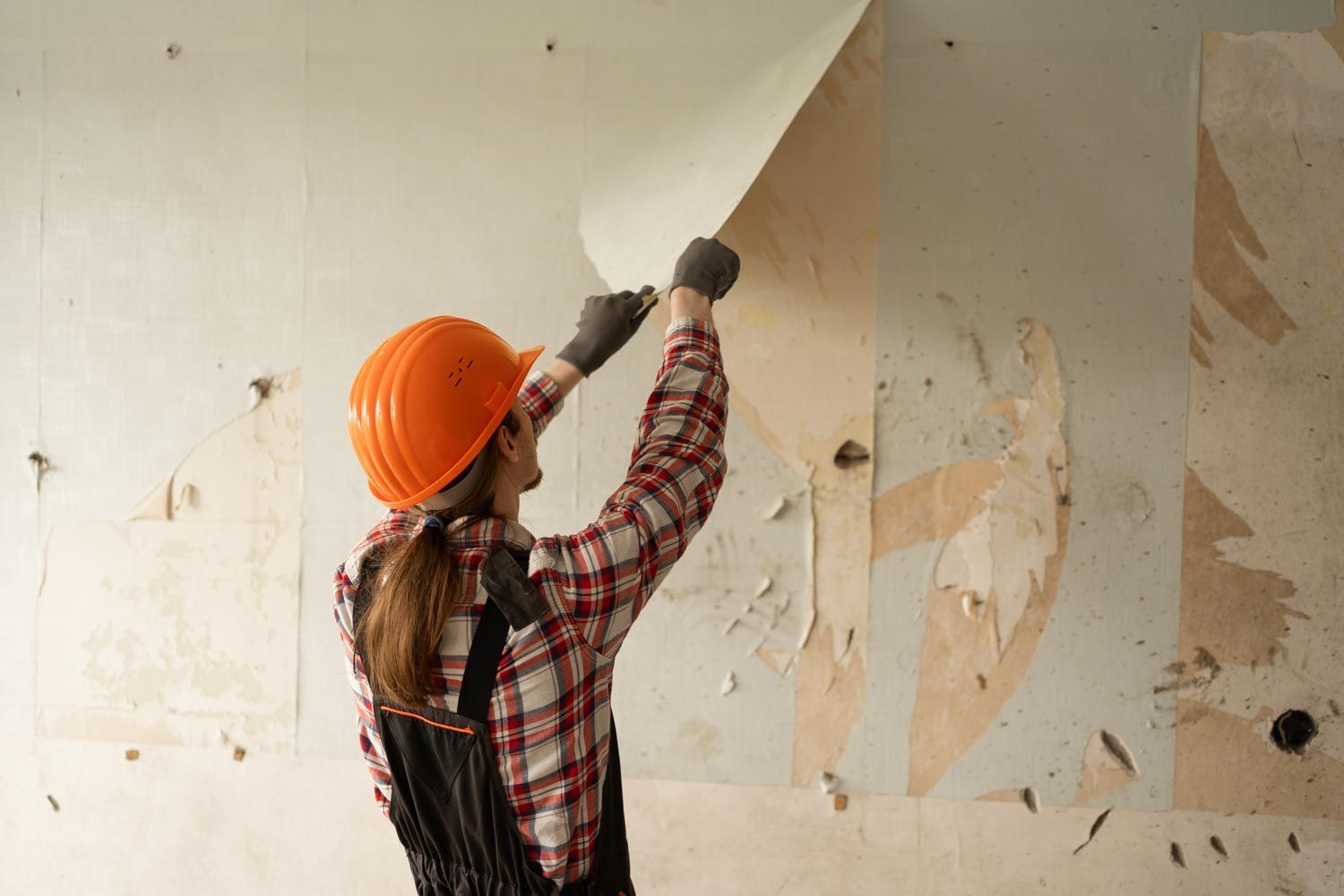 Person in orange hard hat scrapes peeling paint from a wall with a putty knife.