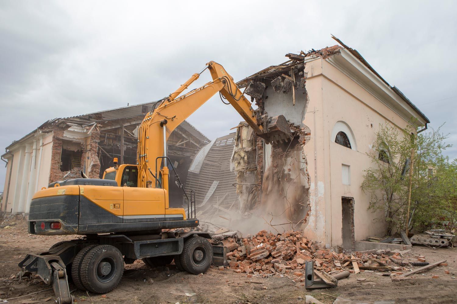 Yellow excavator demolishing a damaged building, with rubble falling around the site