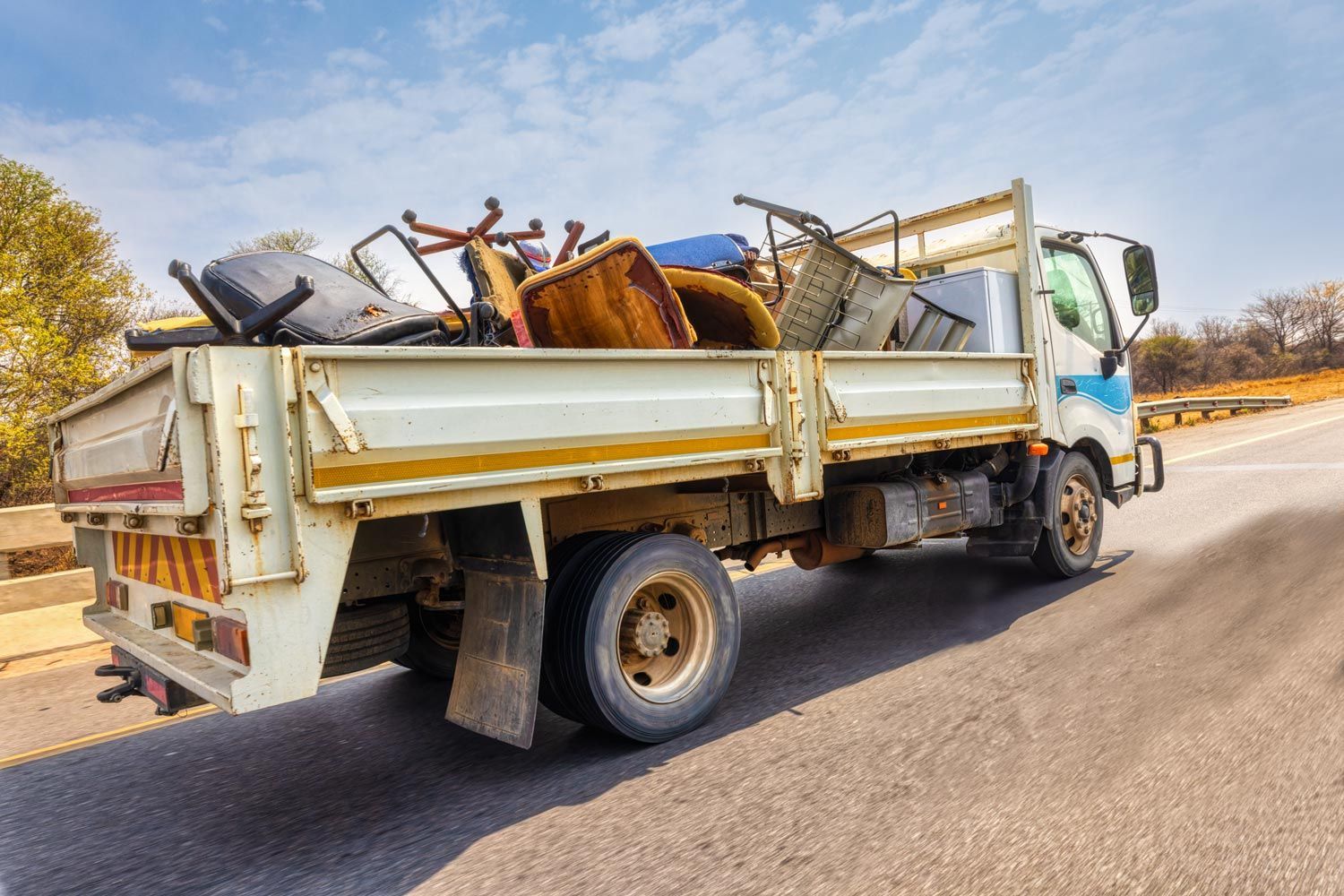 White flatbed truck hauling scrap metal on a roadside in bright daylight