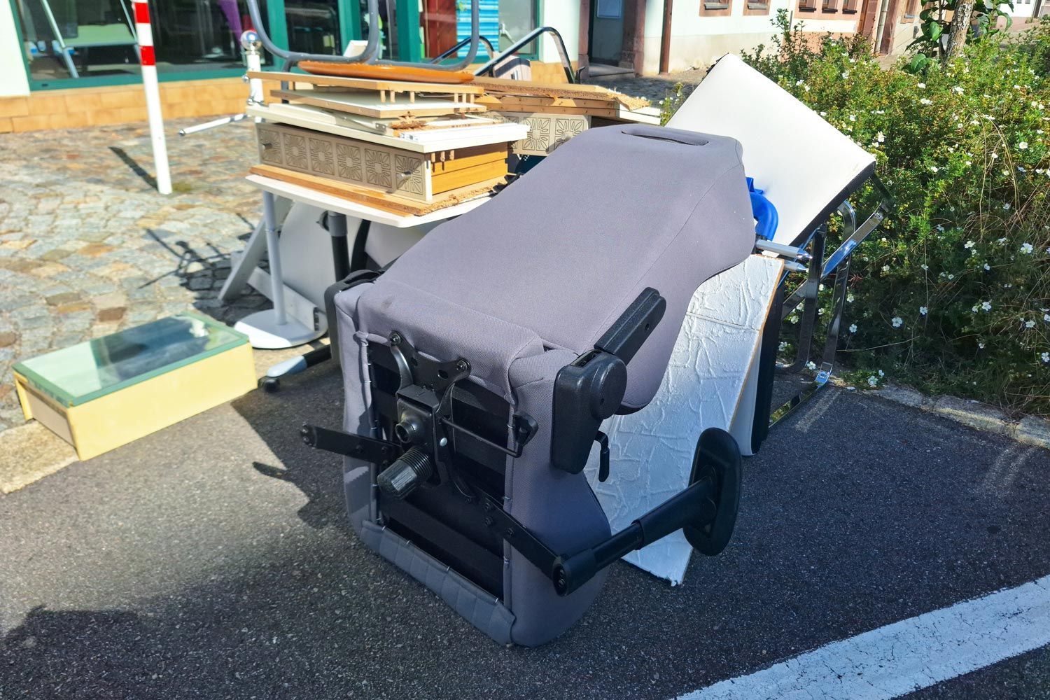 Wheelchair with a gray cover parked outdoors beside a curb and bushes, with furniture in the background