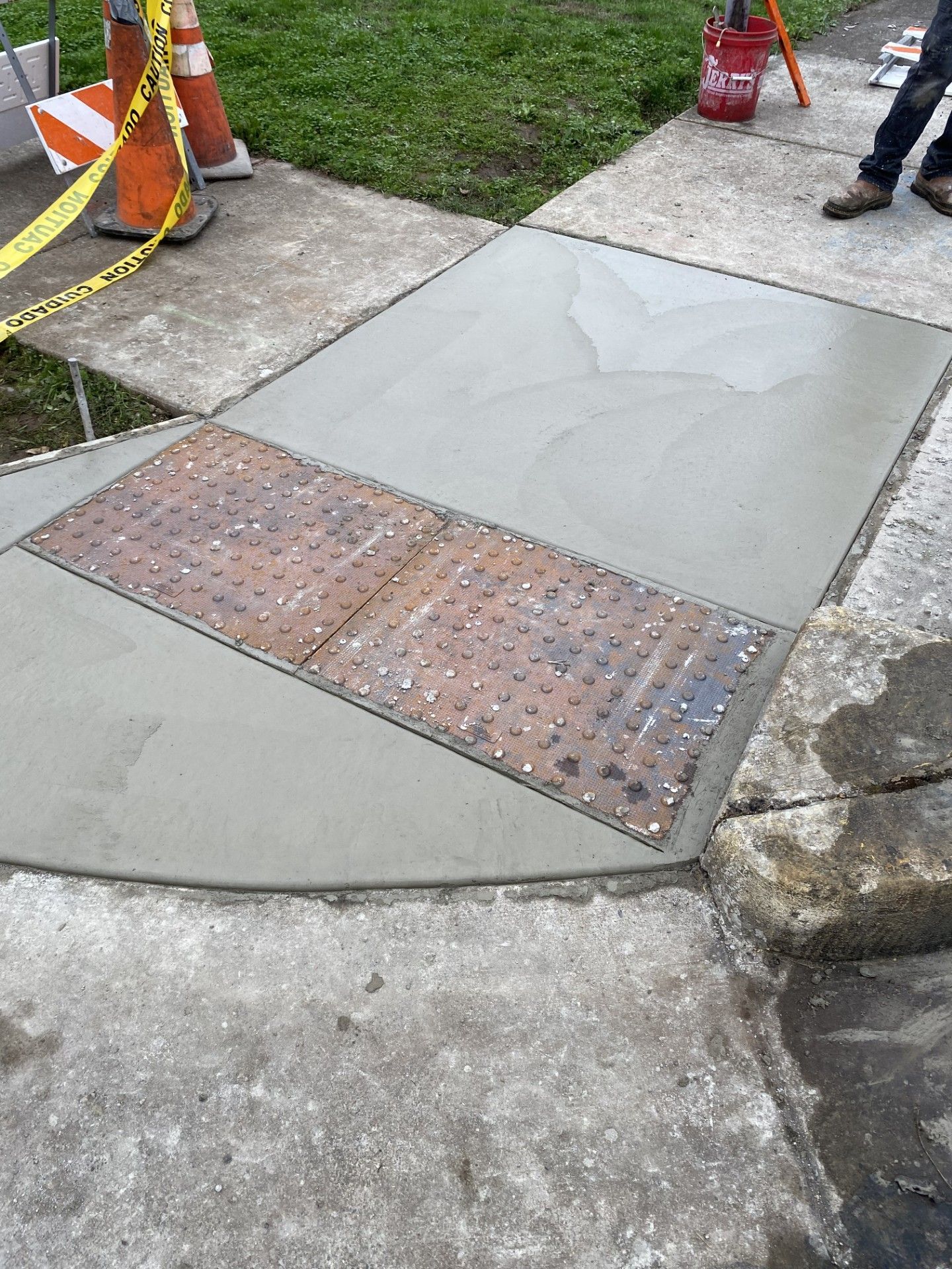Paver stones being laid on a sandy driveway beside a worker’s legs and tools.