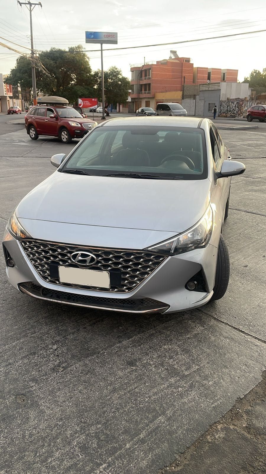 Un Hyundai Accent plateado estacionado en un aparcamiento al aire libre, con un vehículo rojo y edificios al fondo.