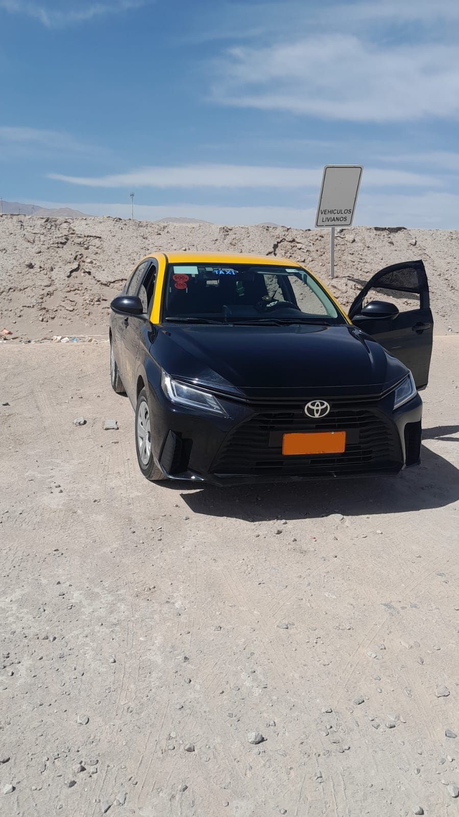 Un taxi Toyota negro con techo amarillo estacionado en un paisaje soleado, árido y desértico, con la puerta abierta.