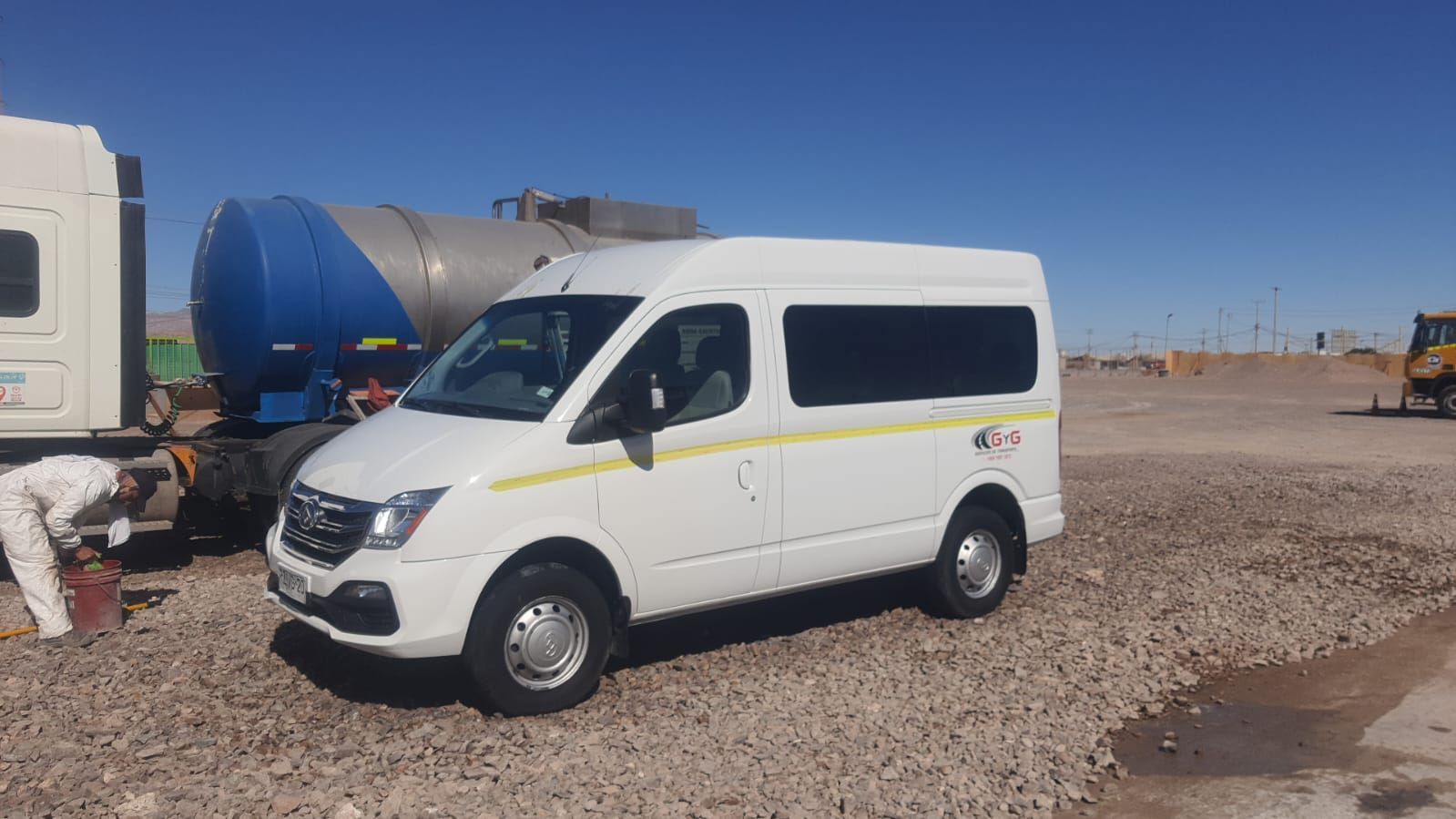Una furgoneta blanca de pasajeros aparcada en un terreno de grava junto a un gran camión