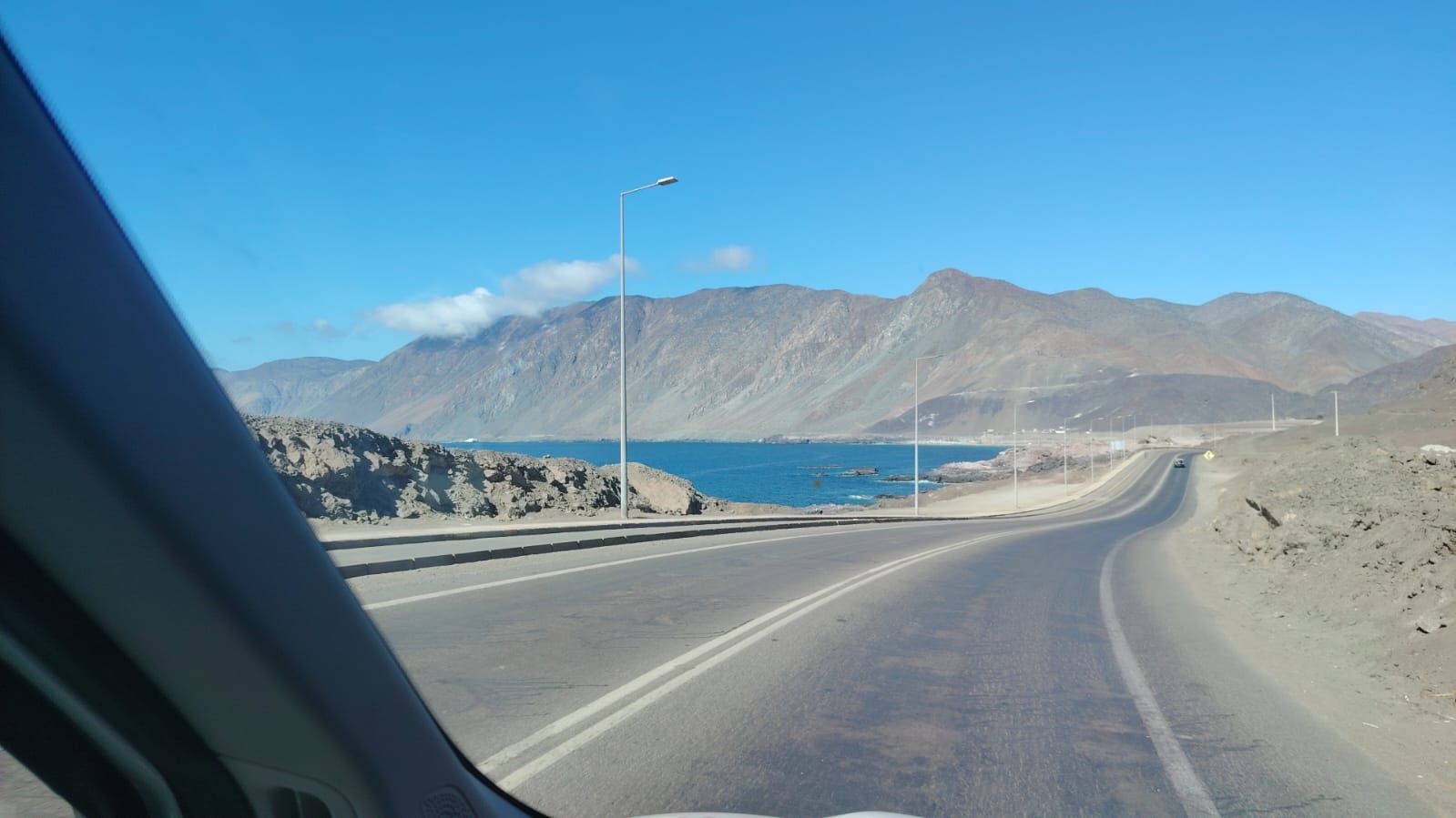 Una carretera asfaltada serpentea a lo largo de la costa, con montañas áridas al fondo, bajo un cielo azul despejado.