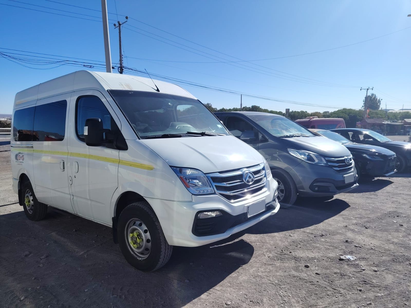 Una furgoneta blanca de pasajeros aparcada en un terreno de tierra junto a una minivan gris y otro vehículo