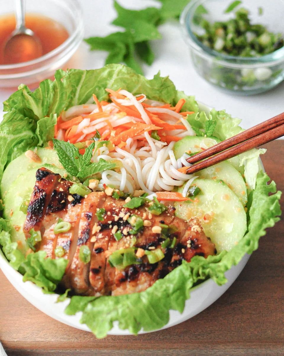 Vietnamese noodle bowl with grilled meat, cucumbers, carrots, herbs, and chopsticks.
