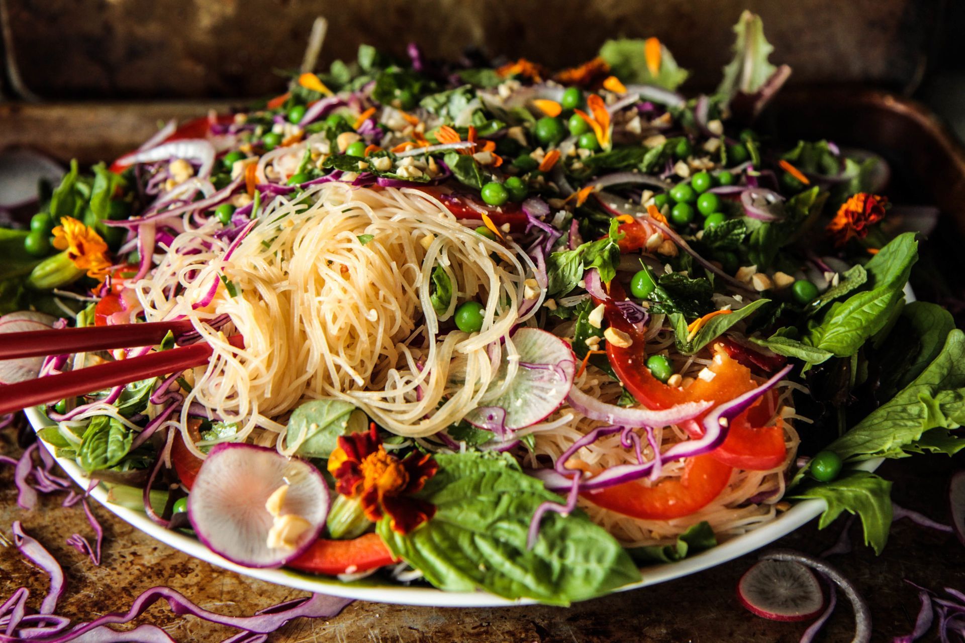 Colorful Asian-style noodle salad with red chopsticks in a white bowl, close-up. Features noodles, vegetables, and herbs.